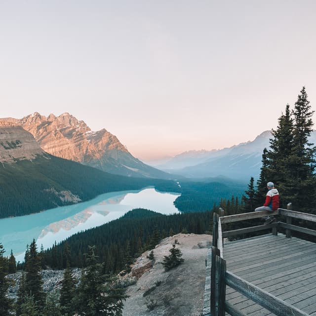 Person sitting on a balcony ledge looking out to a lake surrounded by mountains.